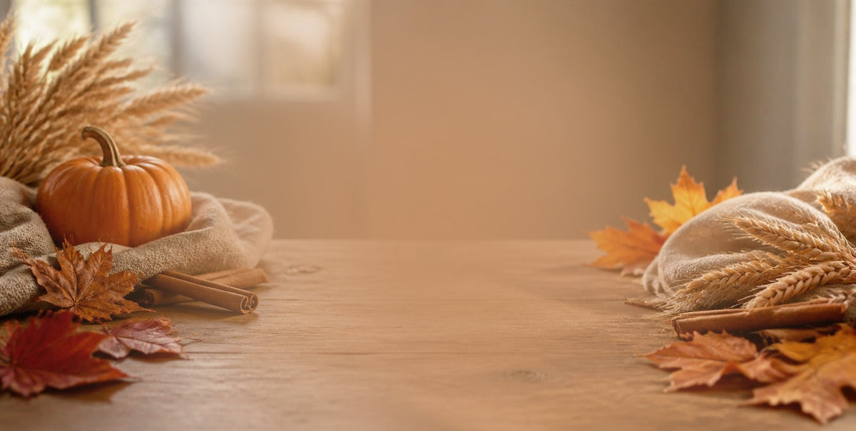 Autumn tabletop decor with a small orange pumpkin, wheat stalks, cinnamon sticks, and fall leaves arranged on a wooden surface, with a softly blurred indoor background.