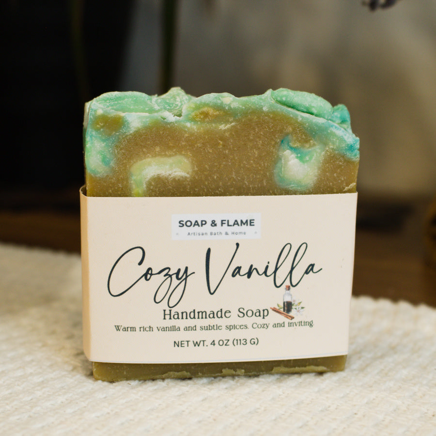 Handmade soap bar with 'Cozy Vanilla' label by Soap & Flame on a textured surface.