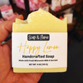 Handcrafted soap bar labeled 'Happy Lemon' by Soap & Flame held in a hand with a store shelf in the background.