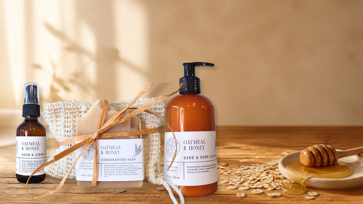 A set of oatmeal and honey hand and foot care products arranged on a wooden surface against a warm beige background. The set includes an amber spray bottle, a wrapped bar of soap tied with a bow, a large amber pump bottle, loose oats, honey, and a honey dipper.