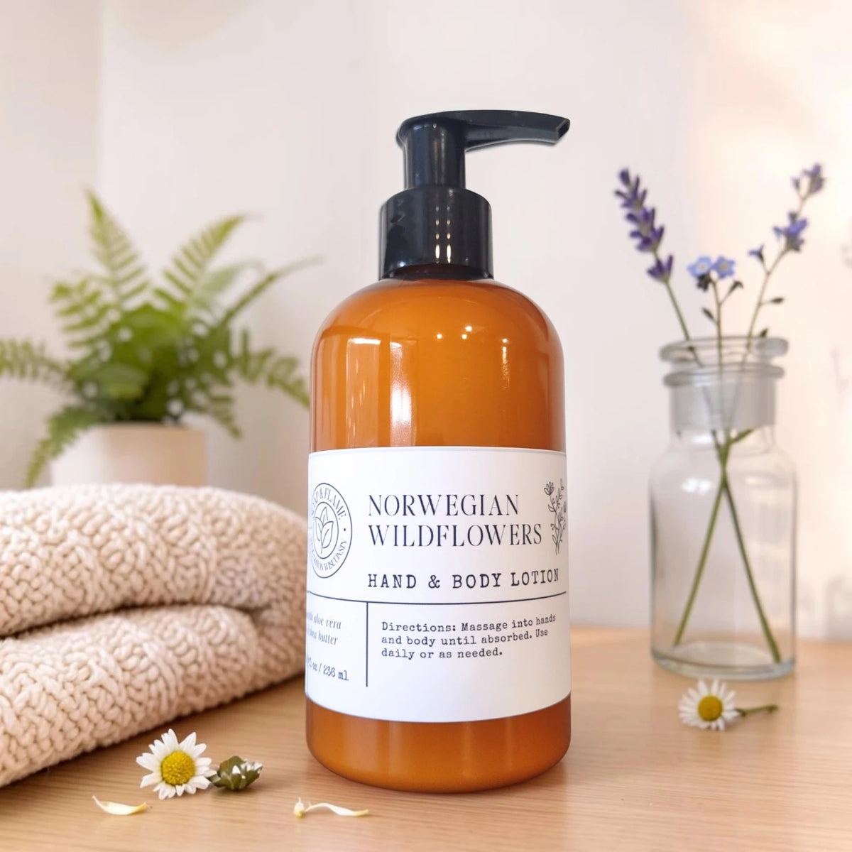 amber bottle of norwegian wildflowers hand and body lotion on a wood surface with a knit blanket folded next to it