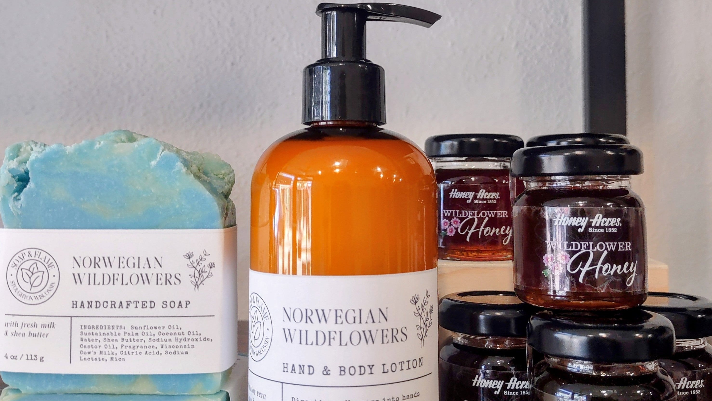 A collection of natural body care products from Norwegian Wildflowers, including a large amber hand and body lotion bottle, a light blue handmade soap bar, and several small jars of honey-flavored lip balm, arranged on a wooden surface against a neutral wall.
