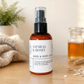 An amber glass spray bottle of Oatmeal & Honey Hand & Body Lotion stands on a wooden surface with a white label. The background features a soft-focus beige wall, a green plant in a pot, a knitted beige cloth