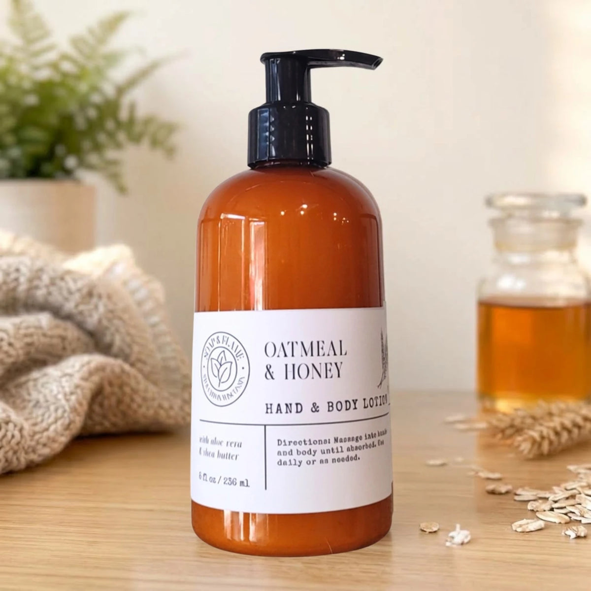 An amber plastic pump bottle of Oatmeal & Honey Hand & Body Lotion set on a wooden surface with a cozy, rustic background featuring a knitted blanket, oats, and a small glass jar.