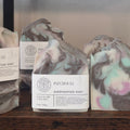 Several handmade soap bars with white labels are arranged on a wooden surface. The center soap is labeled 'PATCHOULI HANDCRAFTED SOAP' and has a marbled swirl pattern in gray, white, black, and pastel pink and mint green.