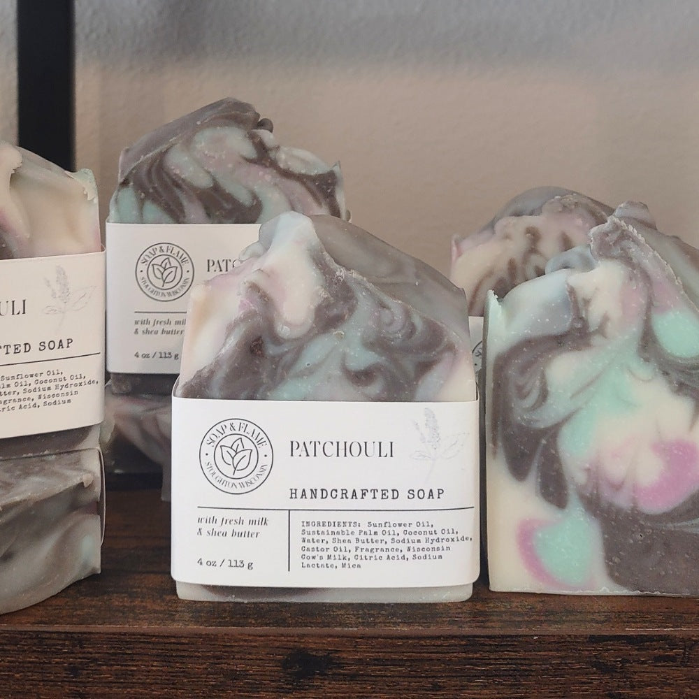 Several handmade soap bars with white labels are arranged on a wooden surface. The center soap is labeled 'PATCHOULI HANDCRAFTED SOAP' and has a marbled swirl pattern in gray, white, black, and pastel pink and mint green.