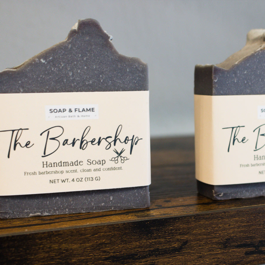 Two bars of handmade soap from Soap & Flame on a wooden surface with a gray background.