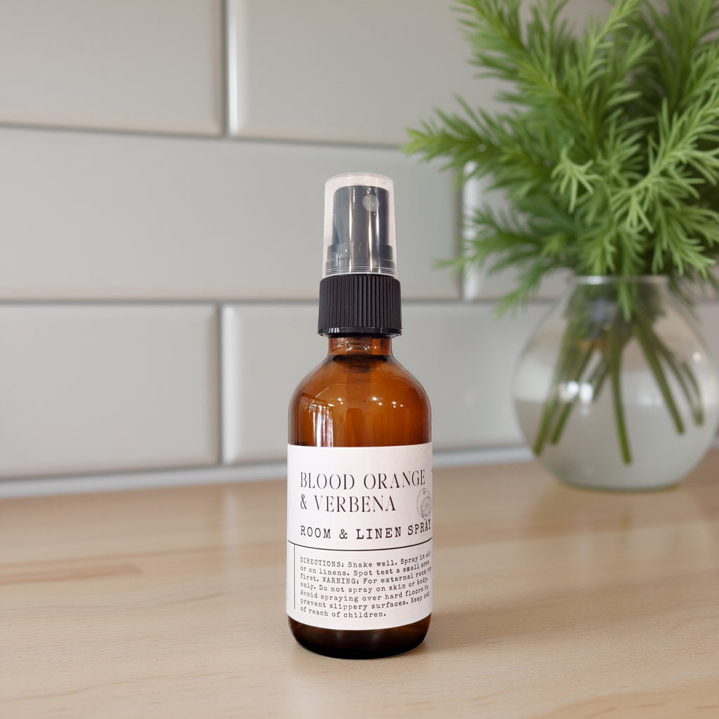 An amber glass room and linen spray bottle with a white label reading 'BLOOD ORANGE & VERBENA' and 'ROOM & LINEN SPRAY,' standing on a light wood surface with a blurred green plant and white tile wall in the background.