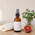 An amber glass room and linen spray bottle with a black spray top and white label reading 'BLOOD ORANGE & VERBENA ROOM & LINEN SPRAY,' styled on a light wood surface with a folded white towel, a halved blood orange, and greenery in the background.