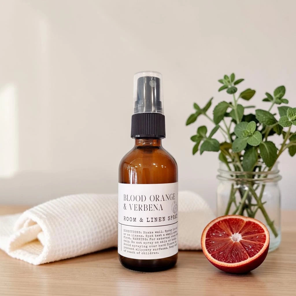 An amber glass room and linen spray bottle with a black spray top and white label reading 'BLOOD ORANGE & VERBENA ROOM & LINEN SPRAY,' styled on a light wood surface with a folded white towel, a halved blood orange, and greenery in the background.