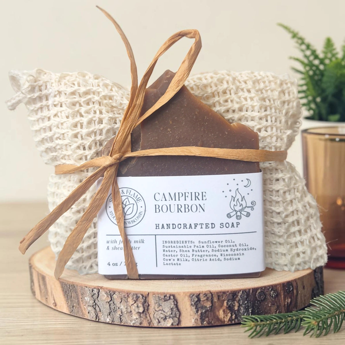 Campfire Bourbon Essentials Gift Set: a soap bar tied with raffia in a natural sisal bag, displayed on a wood slice with a small potted plant, amber glass, and a sprig of pine in the background.