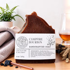 A handcrafted bar of brown soap labeled 'Campfire Bourbon' sits on a wooden surface with a knit blanket, a plant, a glass of amber liquid, and small decorative props around it.
