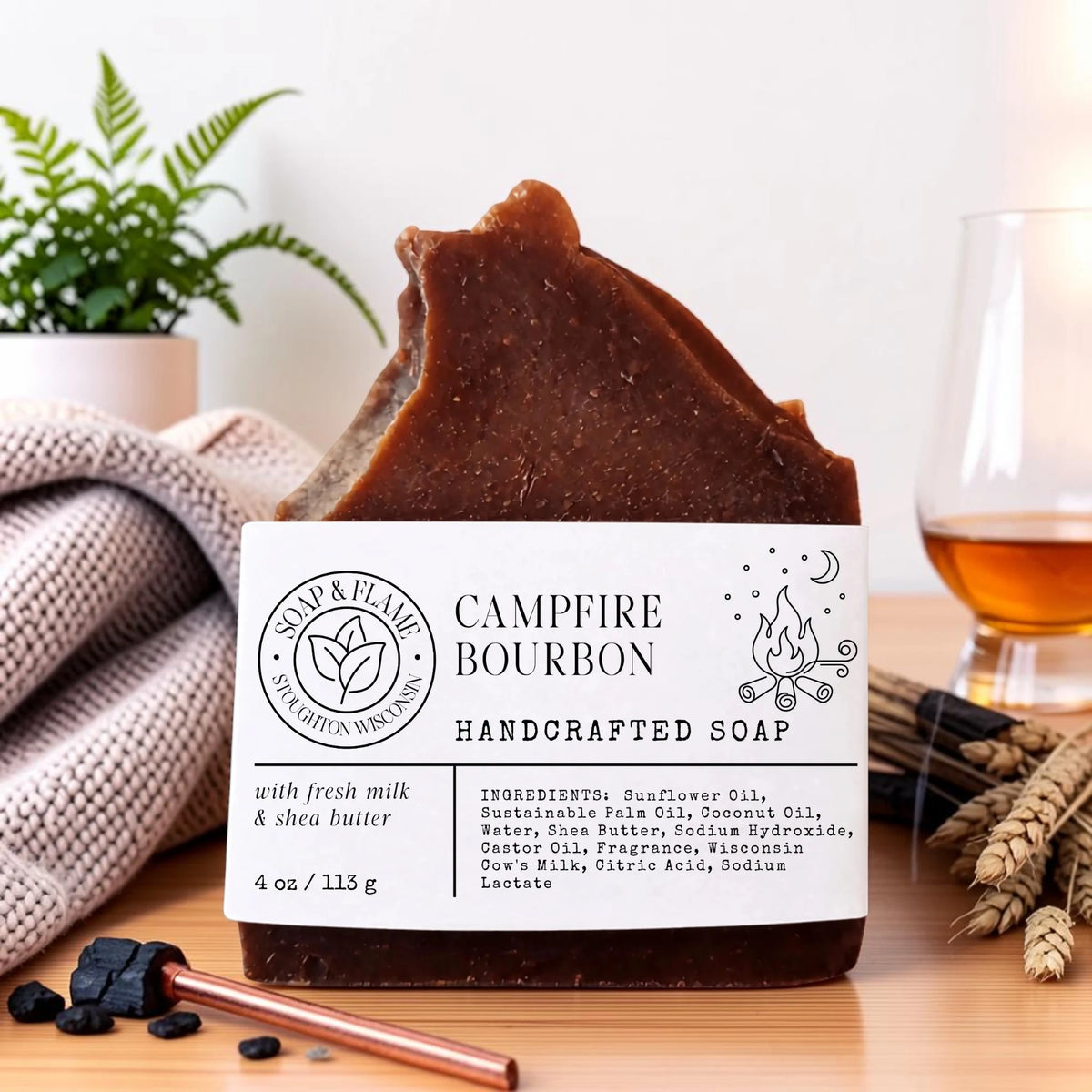 A handcrafted bar of brown soap labeled 'Campfire Bourbon' sits on a wooden surface with a knit blanket, a plant, a glass of amber liquid, and small decorative props around it.