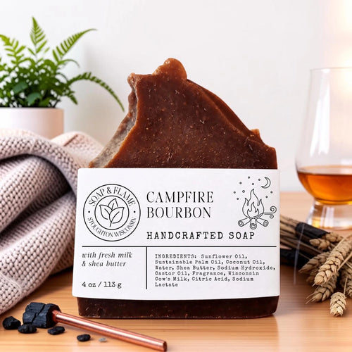A handcrafted bar of brown soap labeled 'Campfire Bourbon' sits on a wooden surface with a knit blanket, a plant, a glass of amber liquid, and small decorative props around it.