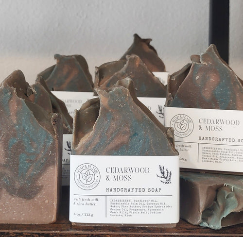 Several rustic handmade soap bars with white labels reading Cedarwood & Moss are arranged on a wooden surface against a light wall.