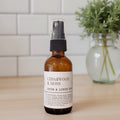 An amber glass spray bottle labeled 'CEDARWOOD & MOSS ROOM & LINEN SPRAY' with a black pump, standing on a light wood surface with a white subway tile wall and a blurred green plant in the background.