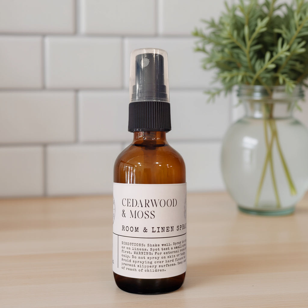 An amber glass spray bottle labeled 'CEDARWOOD & MOSS ROOM & LINEN SPRAY' with a black pump, standing on a light wood surface with a white subway tile wall and a blurred green plant in the background.