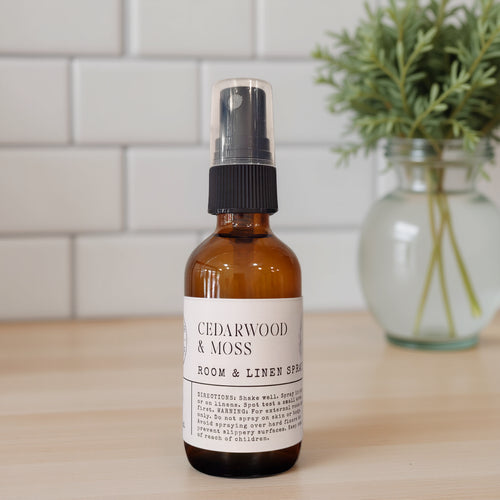 An amber glass spray bottle labeled 'CEDARWOOD & MOSS ROOM & LINEN SPRAY' with a black pump, standing on a light wood surface with a white subway tile wall and a blurred green plant in the background.