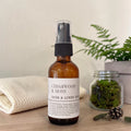 A bottle of Cedarwood & Moss Room & Linen Spray stands on a wooden surface, styled with a folded cream towel, a small jar of moss, and a fern in the background.