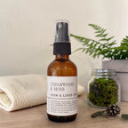 A bottle of Cedarwood & Moss Room & Linen Spray stands on a wooden surface, styled with a folded cream towel, a small jar of moss, and a fern in the background.