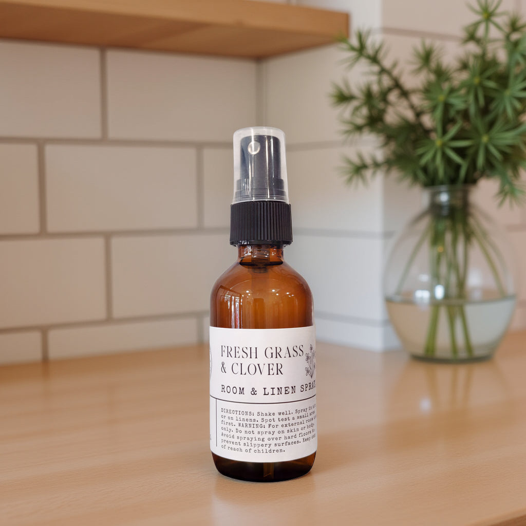 An amber glass room and linen spray bottle with a black spray top and white label reading 'FRESH GRASS & CLOVER ROOM & LINEN SPRAY,' placed on a light wood surface with a tiled wall and a blurred green plant in the background.