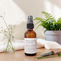 An amber glass room and linen spray bottle with a black spray top and a white label reading 'FRESH GRASS & CLOVER ROOM & LINEN SPRAY,' styled on a tabletop with a folded towel, a small jar of clover blossoms, and a garden tool in the background.
