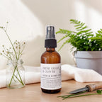 An amber glass room and linen spray bottle with a black spray top and a white label reading 'FRESH GRASS & CLOVER ROOM & LINEN SPRAY,' styled on a tabletop with a folded towel, a small jar of clover blossoms, and a garden tool in the background.