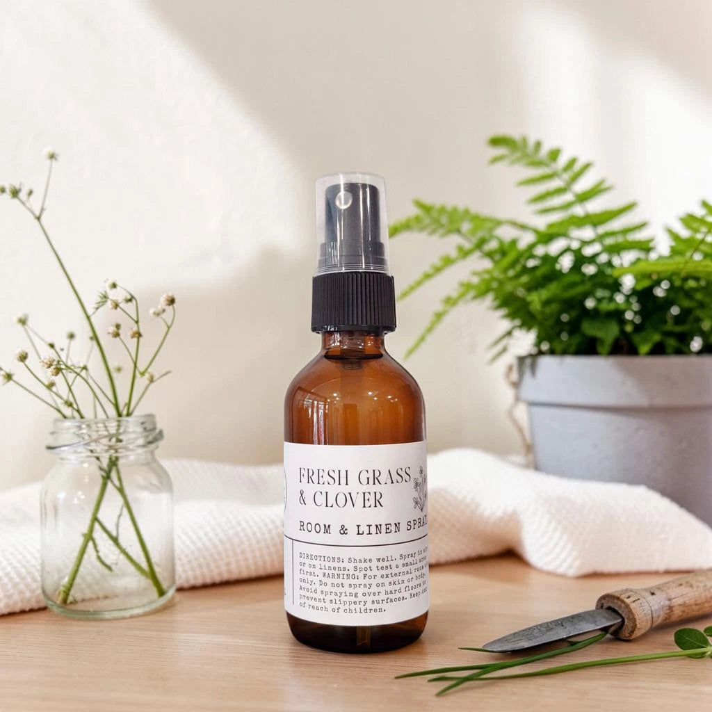 An amber glass room and linen spray bottle with a black spray top and a white label reading 'FRESH GRASS & CLOVER ROOM & LINEN SPRAY,' styled on a tabletop with a folded towel, a small jar of clover blossoms, and a garden tool in the background.