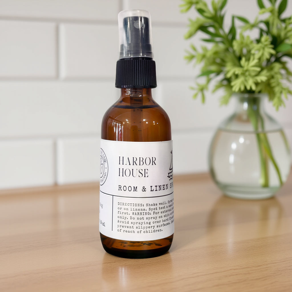 An amber glass room and linen spray bottle with a black spray top and white label reading HARBOR HOUSE, placed on a light wood surface with a blurred green plant and white tile wall in the background.