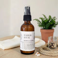 An amber glass room and linen spray bottle with a black spray top and a white label reading 'HARBOR HOUSE ROOM & LINEN', placed on a wooden surface with a folded white towel, a potted fern, and a small jar in the background.