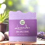 Lavender sea salt soap with 'Soap & Flame' branding on a wooden surface with blurred greenery in the background.