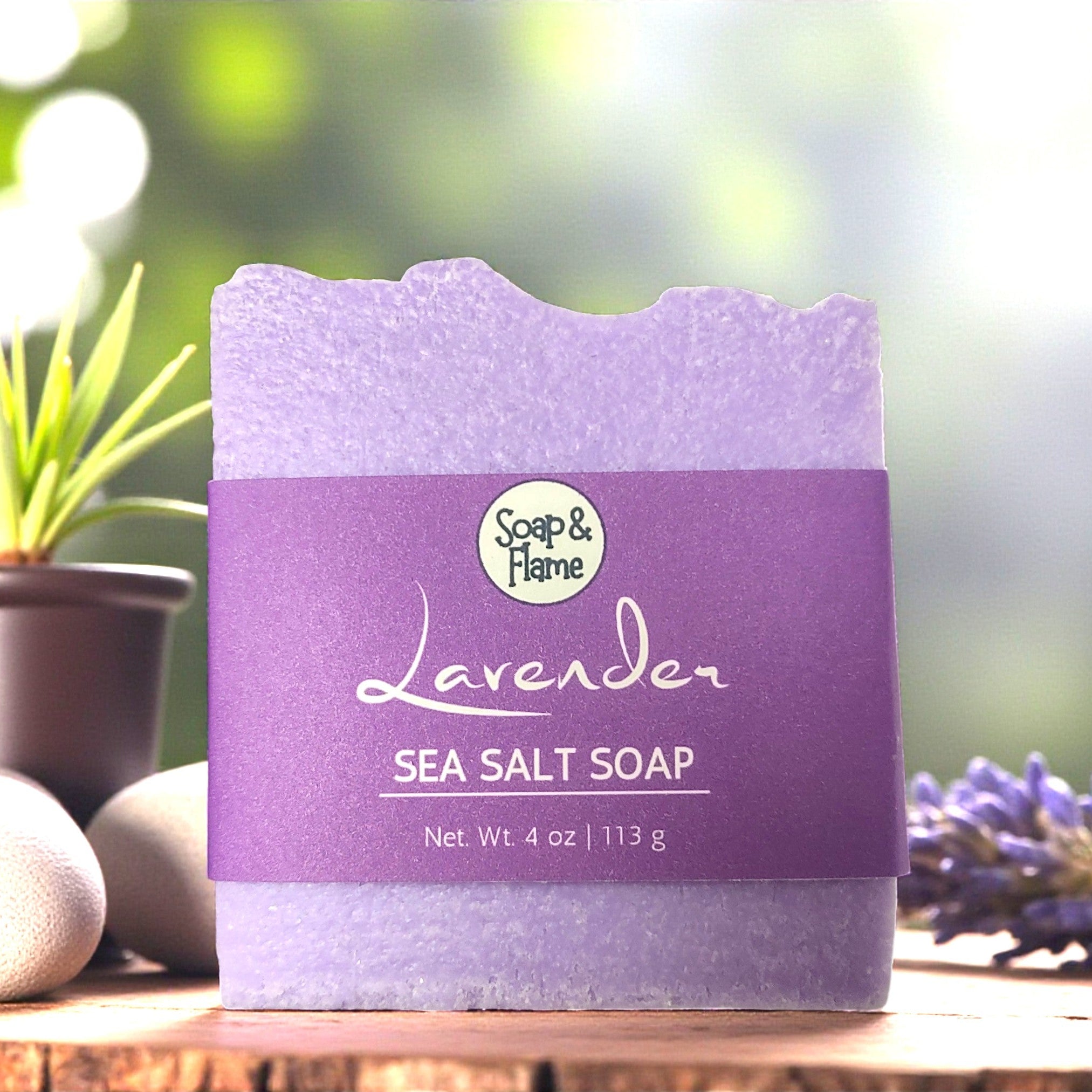 Lavender sea salt soap with 'Soap & Flame' branding on a wooden surface with blurred greenery in the background.