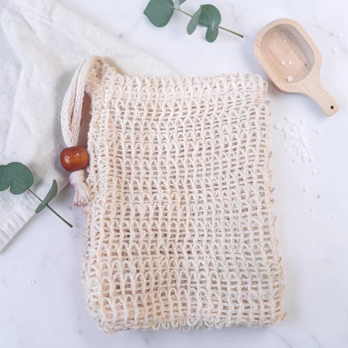 Natural Sisal Soap Bag: a cream sisal drawstring pouch in a flat lay with a wooden scoop, eucalyptus sprigs, folded cloth, and scattered sea salt.