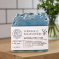 A handmade bar of soap labeled 'Norwegian Wildflowers Handcrafted Soap' by Soap & Flame, resting on a wooden surface with a blurred kitchen background.