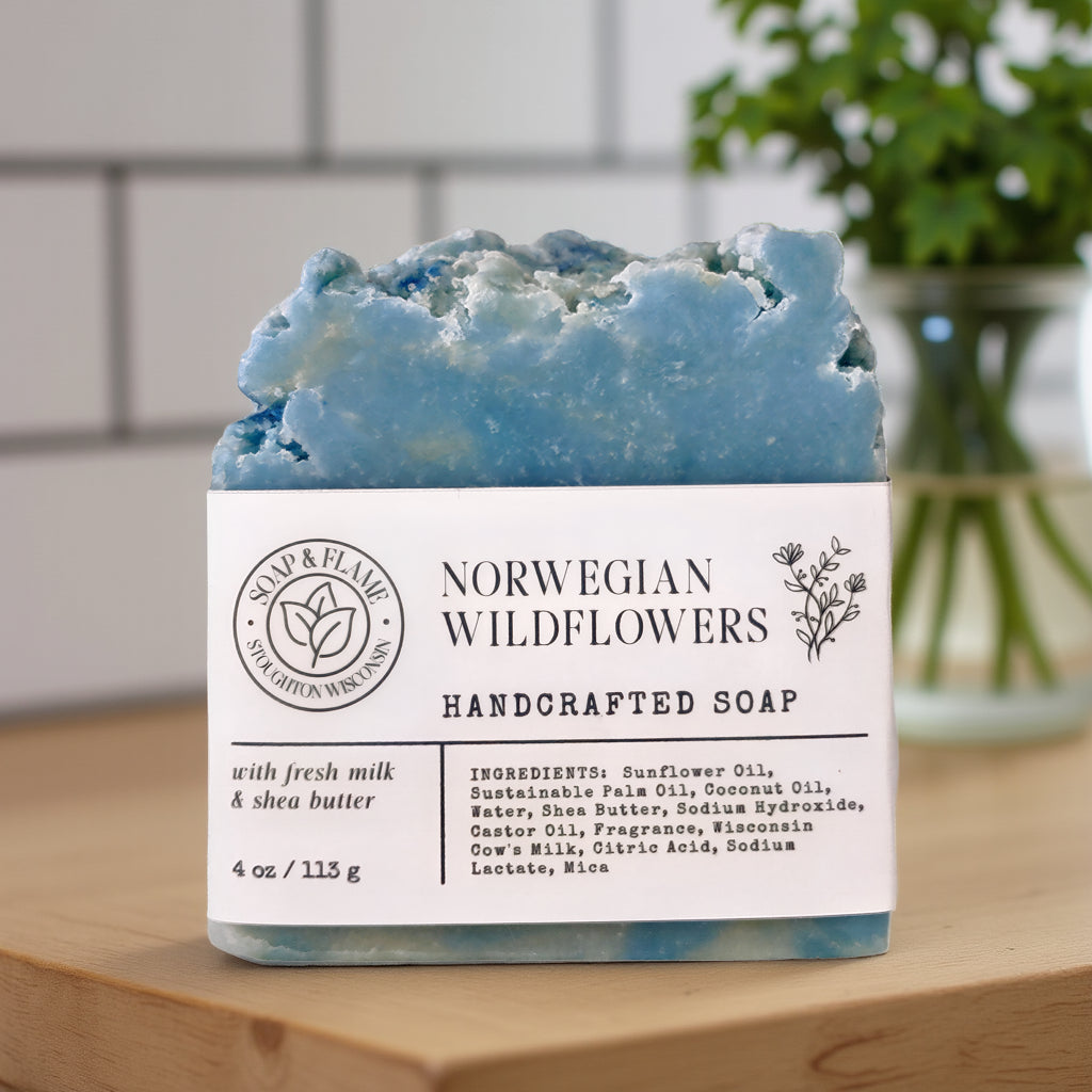 A handmade bar of soap labeled 'Norwegian Wildflowers Handcrafted Soap' by Soap & Flame, resting on a wooden surface with a blurred kitchen background.