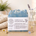 A single bar of handmade soap stands upright on a wooden surface, wrapped in a white paper label that reads 'SOAP & FLAME' and 'NORWEGIAN WILDFLOWERS HANDCRAFTED SOAP.' The soap itself is a pale icy blue.