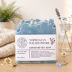 A single bar of handmade soap stands upright on a wooden surface, wrapped in a white paper label that reads 'SOAP & FLAME' and 'NORWEGIAN WILDFLOWERS HANDCRAFTED SOAP.' The soap itself is a pale icy blue.