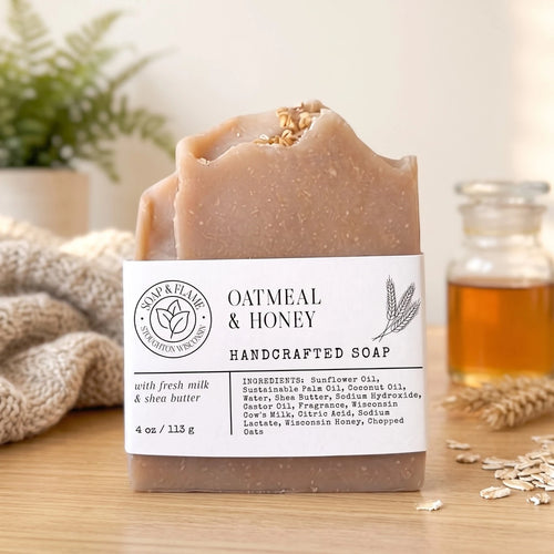 A handcrafted oatmeal and honey soap bar with a white label, displayed on a wooden surface with a cozy background of a knitted blanket, a small plant, and a glass bottle of honey.
