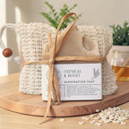 Oatmeal & Honey Essentials Gift Set: an oat-flecked soap bar tied with raffia in a natural sisal bag, displayed on a round wood board with scattered oats and a small honey jar.