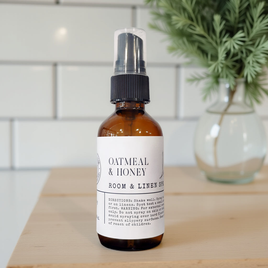 An amber glass spray bottle labeled 'Oatmeal & Honey Room & Linen Spray' with a black spray top, standing on a light wood surface with a blurred white tile wall and green plant in the background.
