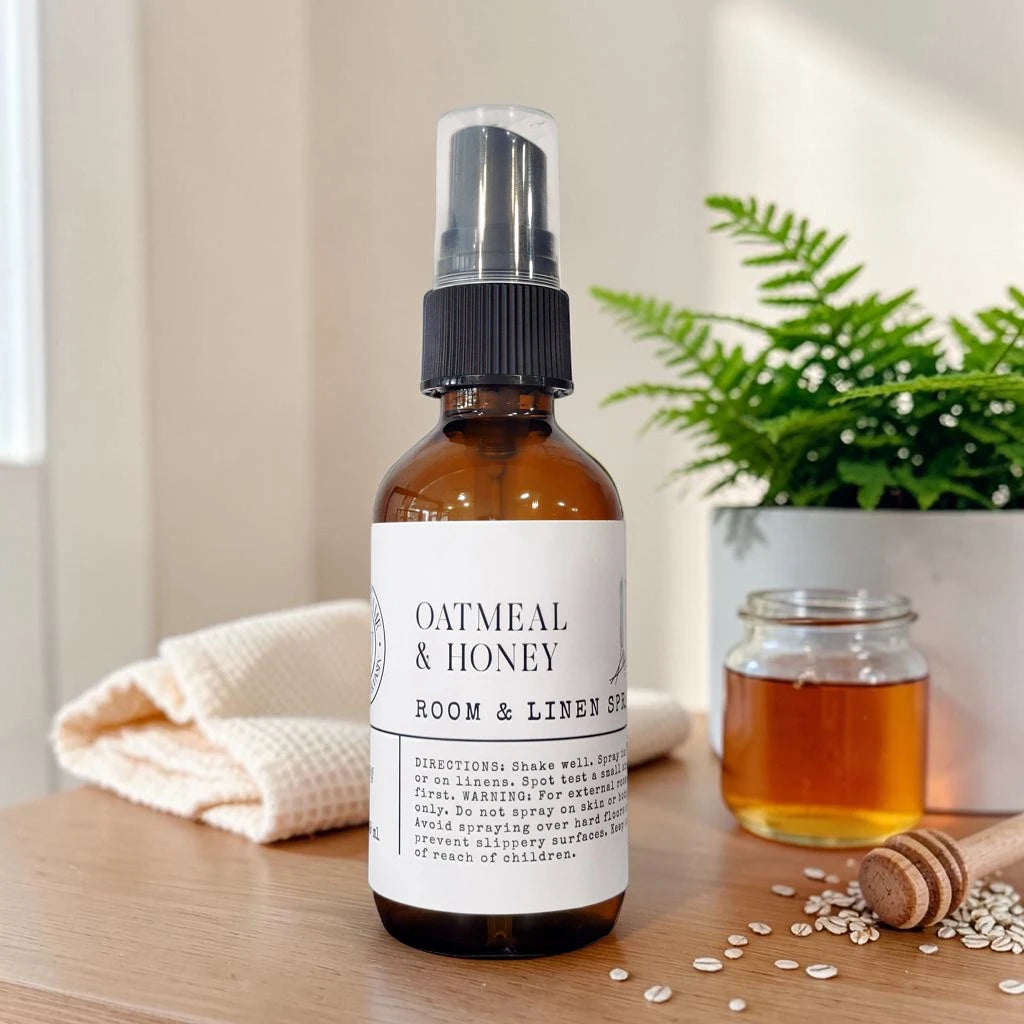An amber glass room spray bottle labeled 'Oatmeal & Honey Room & Linen Spray' stands on a wooden surface with a folded cream towel, a honey jar, oats, and a green plant in the background.