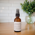 An amber glass room spray bottle with a black spray top and a white label reading 'ORCHID & PINK GRAPEFRUIT ROOM & LINEN SPRAY', standing on a wooden surface with a blurred white tile wall and green plant in the background.