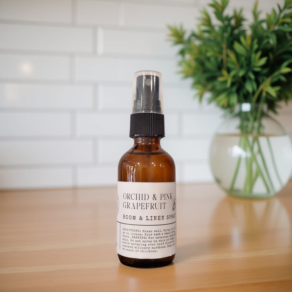 An amber glass room spray bottle with a black spray top and a white label reading 'ORCHID & PINK GRAPEFRUIT ROOM & LINEN SPRAY', standing on a wooden surface with a blurred white tile wall and green plant in the background.