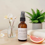 An amber glass room spray bottle labeled 'ORCHID & PINK GRAPEFRUIT ROOM & LINEN SPRAY' stands on a light wood surface with a grapefruit slice, a small glass jar with white flowers, and a potted fern in the background.