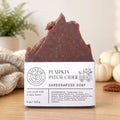 Handcrafted pumpkin patch cider soap bar with a white paper label, displayed on a wooden surface with a cozy background of a knitted blanket, potted fern, and small pumpkins.