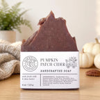 Handcrafted pumpkin patch cider soap bar with a white paper label, displayed on a wooden surface with a cozy background of a knitted blanket, potted fern, and small pumpkins.