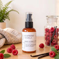 A bottle of Raspberry Vanilla Hand & Body Lotion stands on a wooden surface, surrounded by fresh raspberries, vanilla beans, and a knitted blanket, with a glass jar of raspberries in the background.