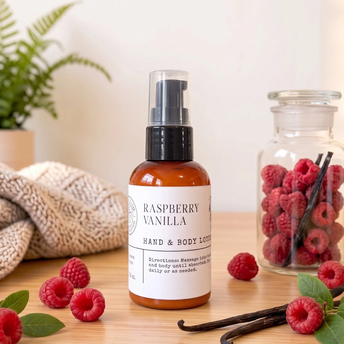 A bottle of Raspberry Vanilla Hand & Body Lotion stands on a wooden surface, surrounded by fresh raspberries, vanilla beans, and a knitted blanket, with a glass jar of raspberries in the background.