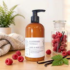 A bottle of Raspberry Vanilla hand and body lotion stands on a wooden surface, surrounded by raspberries, vanilla beans, and a small jar of raspberries. A folded knit blanket and a potted fern are in the background, creating a styled, natural product presentation.