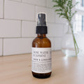 An amber glass room spray bottle with a black spray top and a white label reading 'ROSE WATER & JASMINE ROOM & LINEN SPRAY', standing on a light wood surface with a white tiled wall and a blurred glass vase in the background.